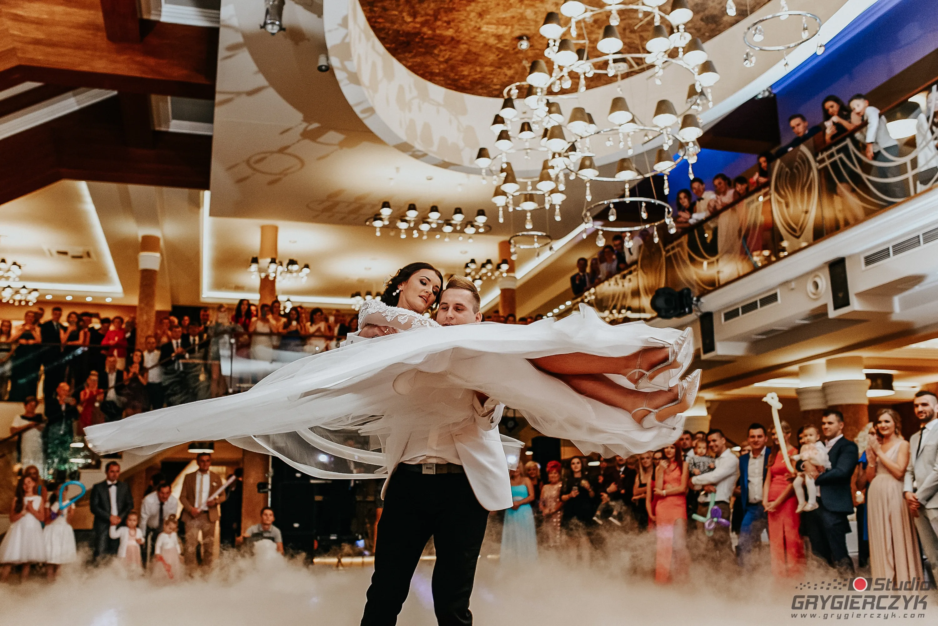 para mloda w dynamicznym tancu slubnym z falujaca suknia trzymana na rekach mlodego pana w nowoczesnej sali slubnej w otoczeniu gosci weselnych patrzacyhc z podziwem na wirujace sylwetki. fotograf grygierczyk lukasz .