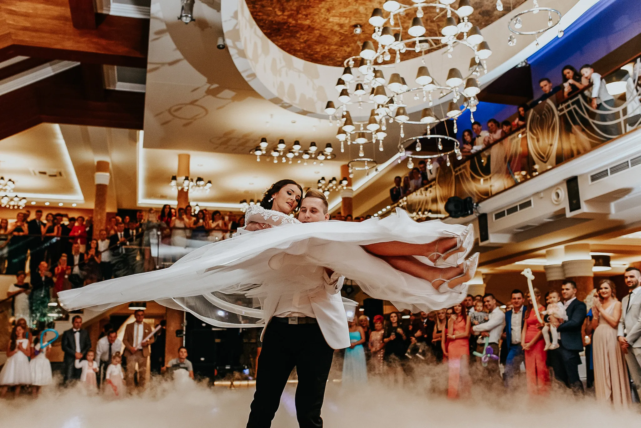 mlody pan wiruje w pierwszym tancu z mloda pania trzymajac ja na rekach z falujaca w rytm muzyki suknia slubna