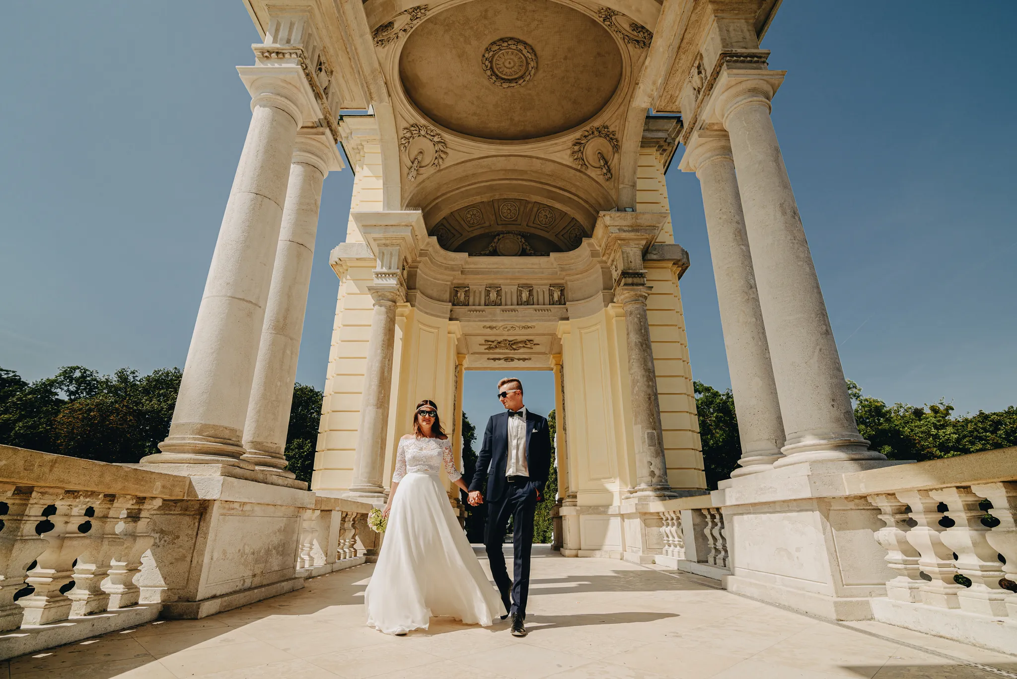 para mloda spaceruje w  museum glorietta w pieknym architektonicznie budynku trzymajac sie za rece .pani mloda ma w zwiewnej sukience z wiankiem na glowie a pan mlody w granatowym garniturze. oboje malzonkow posiada zalozone okulary sloneczne