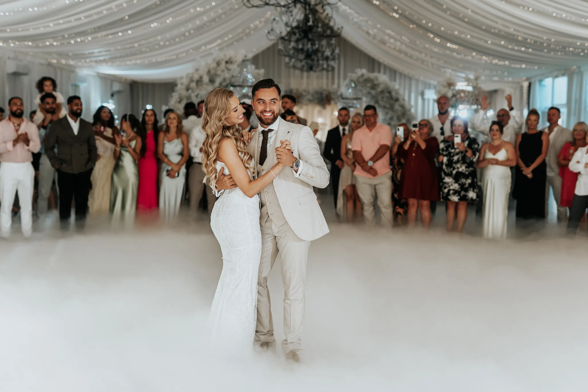 radosna para mloda podczas pierwszego tanca zatopiona w chmurach. Mloda pani ubrana w obcisla sukienke z rozpuszczonymi blond wlosami ukladajace sie efektowne loki. pan mlody ubrany w nowoczesny bezowy garnitur z bardzo dobrze przycieta broda o ciemnej karnacji. hotel bajka opolskie